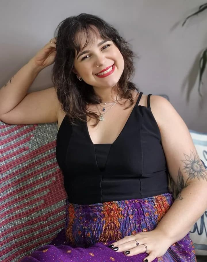 Ariane Aleixo sentada no sofá, sorrindo, com a mão no cabelo.