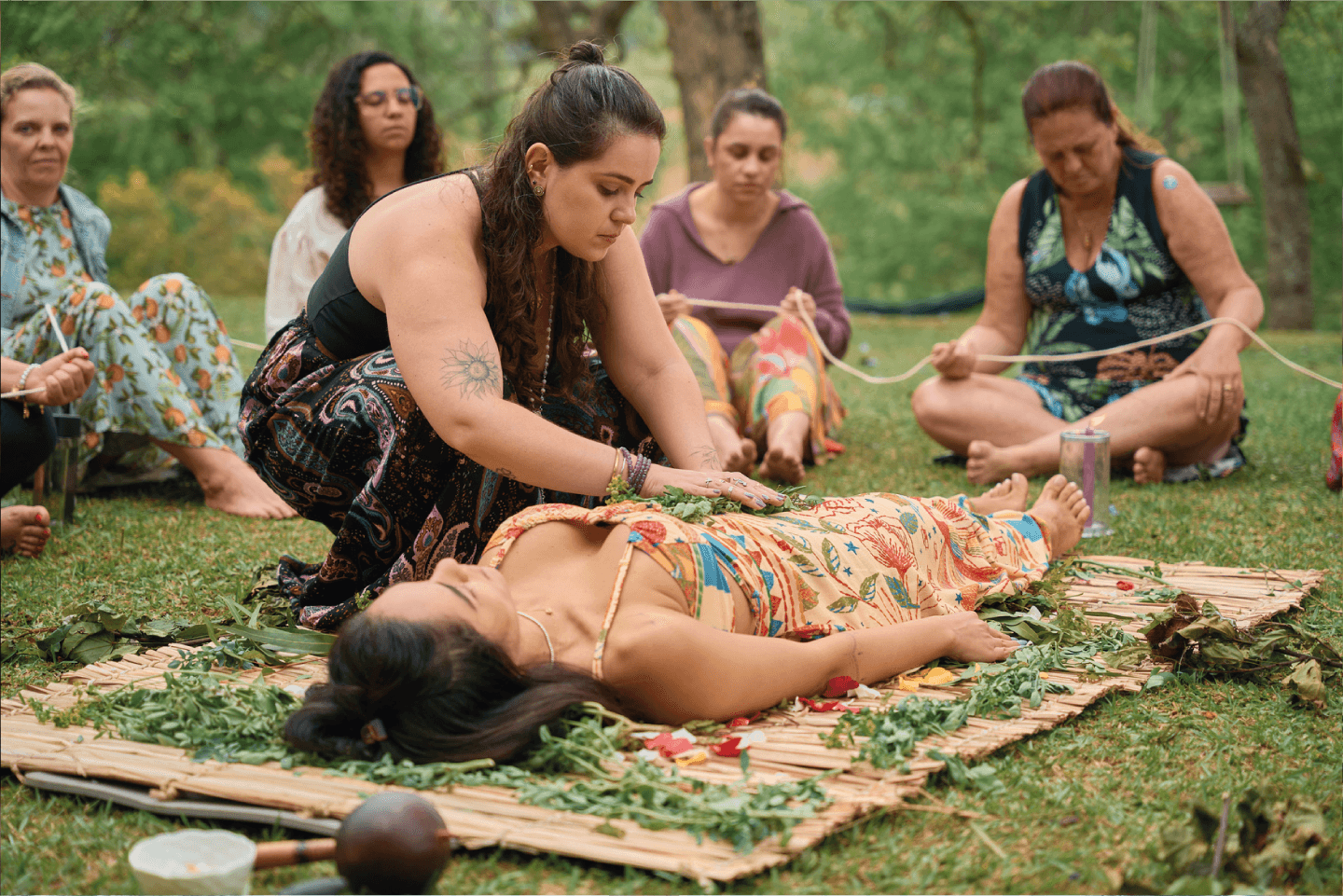 Ariane executando ritual em campo aberto, no centro de um circulo de mulheres sentadas. Ritual ocorrendo em cima de uma esteira de ervas.