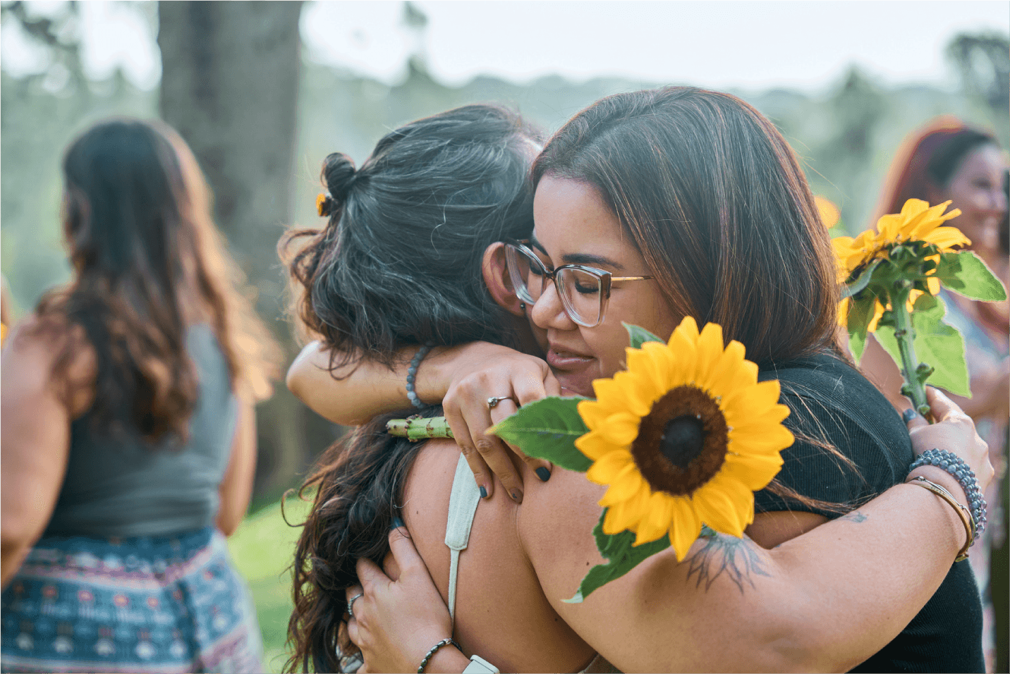 Mulheres a céu aberto se abraçando enquanto seguram um girassol.