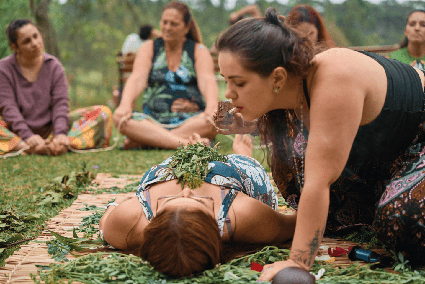 Ariane em um campo aberto, no centro de uma roda de mulheres, executando um ritual em uma moça numa esteira de ervas.